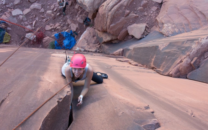 a rock climber smiles up at the camera while navigating a crag on red rocks